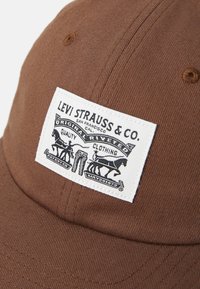 Căciulă din bumbac maronie cu o margine curbă și o etichetă albă cu grafice și text negru pentru Levi Strauss & Co.