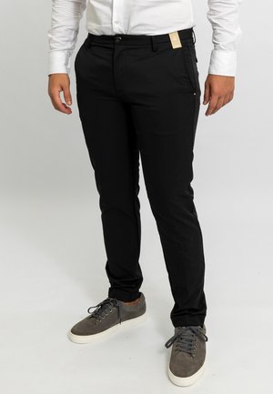 Uomo che indossa pantaloni neri slim fit con cartellino del prezzo, camicia bianca a maniche lunghe e sneakers in suede grigio con suola bianca su sfondo bianco.