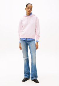 Donna che indossa un hoodie rosa chiaro sopra una camicia a righe blu, abbinato a jeans flare blu chiaro e scarpe nere, in piedi su uno sfondo bianco.