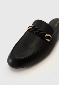 Black leather slip-on shoe featuring a smooth surface, pointed toe, and gold and black chain hardware detail across the vamp.