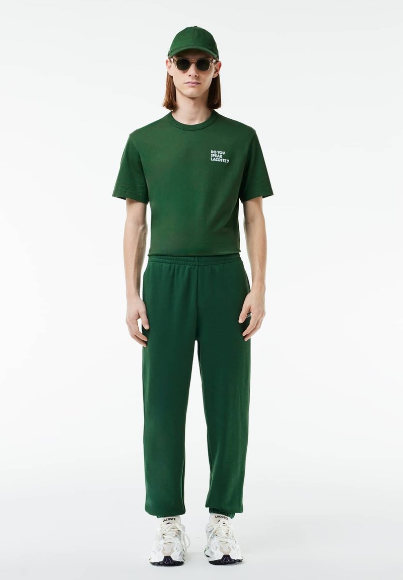 Zielona koszulka bawełniana z napisem "CZY MÓWISZ LACOSTE?", pasujące zielone dresy i białe trampki z kolorowymi akcentami.