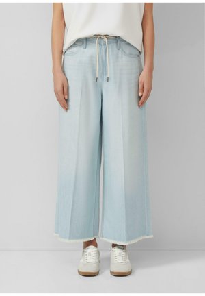 Hellblaue, weit geschnittene, verkürzte Jeans mit rohen Säumen, geschnürter Taille, getragen mit weißen Sneakers und einem in die Hose gesteckten weißen Oberteil.