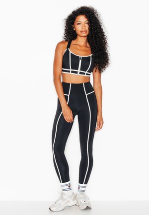 Donna con lunghi capelli ricci che indossa un top sportivo nero e leggings con bordo bianco, calzini bianchi e sneakers bianchi, in piedi su sfondo bianco.