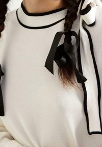 Pull en tricot blanc avec des accents noirs le long du col et des côtés. Il comporte un large ruban noir noué à l'épaule. Texture lisse.