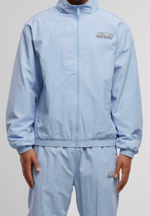 Homme portant une veste zippée bleu clair et un pantalon assorti avec le logo "Kani Sport" sur la poitrine et la cuisse, debout, les bras détendus.