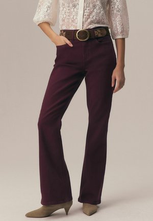 Femme portant un pantalon évasé violet foncé, une ceinture marron avec une boucle ronde, des chaussures à talons beiges et un chemisier en dentelle blanche à manches trois quarts.