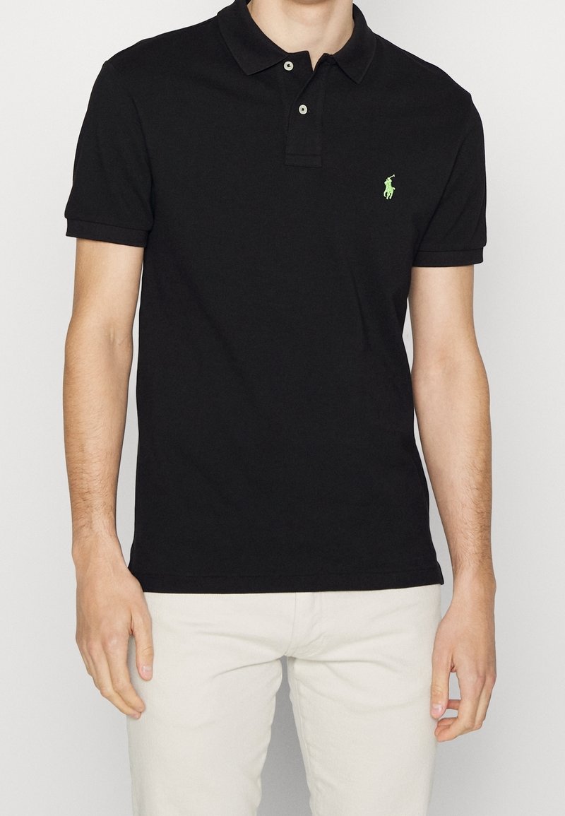 Polo-shirt en coton noir à manches courtes, avec une patte de boutonnage à deux boutons et un logo brodé vert sur la poitrine gauche. Col et poignets en tricot plat.