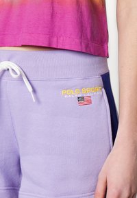 Person som bär lila Polo Sport-shorts med vit dragsko och broderad amerikansk flagga, ihop med en rosa topp.
