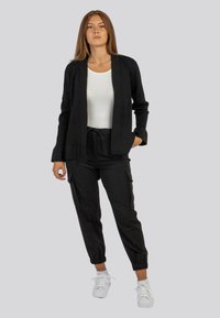 Cardigan aperto nero su un top bianco, abbinato a pantaloni jogger neri con polsini elasticizzati e tasche laterali. Sneakers bianche.