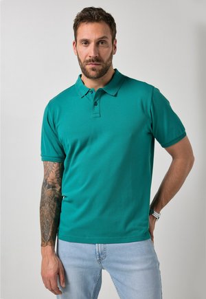 Mężczyzna z brodą i wytatuowanym ramieniem, ubrany w turkusową koszulkę polo i jasnoniebieskie dżinsy, stojący na tle jednolitego szarego tła.