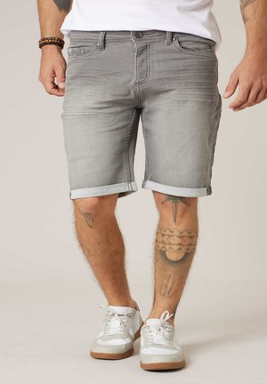 Homme portant un short en denim gris clair retroussé, des baskets blanches et des bracelets en perles, avec des tatouages visibles sur les jambes.