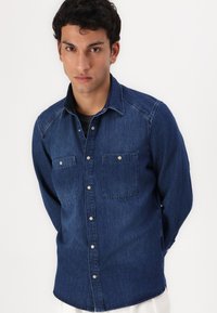 Camisa de mezclilla en azul oscuro con botones en la parte delantera, dos bolsillos en el pecho y un cuello puntiagudo. Presenta una textura suave y un dobladillo ligeramente curvado.