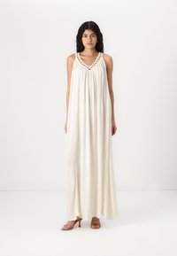 Longue robe maxi blanche avec un décolleté en V, détails tressés le long du cou et tissu fluide pour une coupe ample. Portée avec des talons bruns à brides.