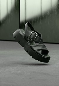 Black platform sandals with cross-strap design, made of smooth leather. Features a metallic buckle and a chunky, ridged sole.
