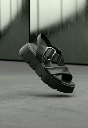 Black platform sandals with cross-strap design, made of smooth leather. Features a metallic buckle and a chunky, ridged sole.