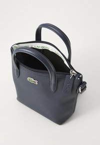 Borsa sintetica blu navy con doppi manici, chiusura con zip e logo Lacoste a contrasto; texture liscia e forma rettangolare compatta.