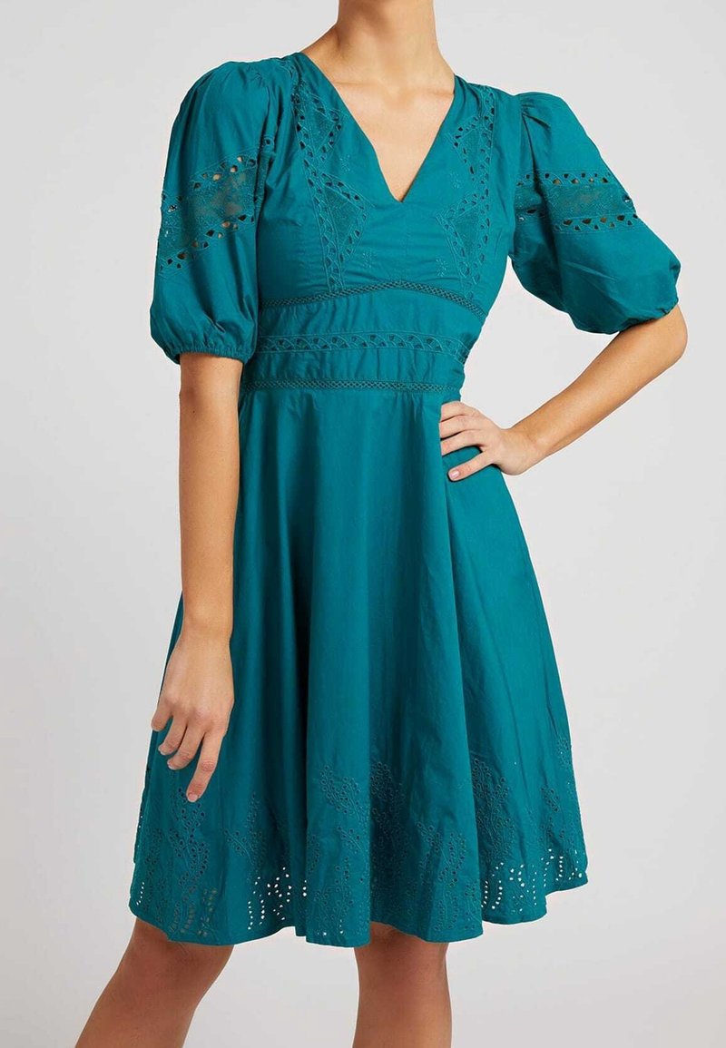 Robe turquoise avec manches bouffantes, col en V et détails complexes de dentelle découpée sur les manches, la taille et l'ourlet, portée avec la main sur la hanche.