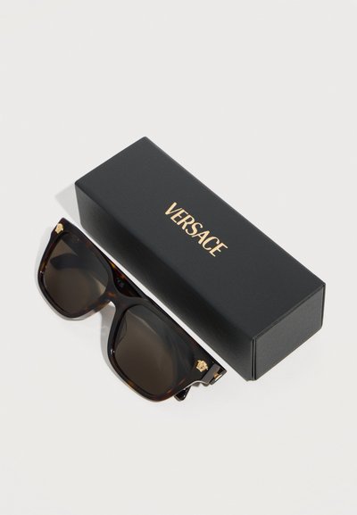 Μαύρα ορθογώνια γυαλιά ηλίου Versace με χρυσό λογότυπο Μέδουσας στα πλαϊνά, δίπλα σε μαύρη θήκη Versace.
