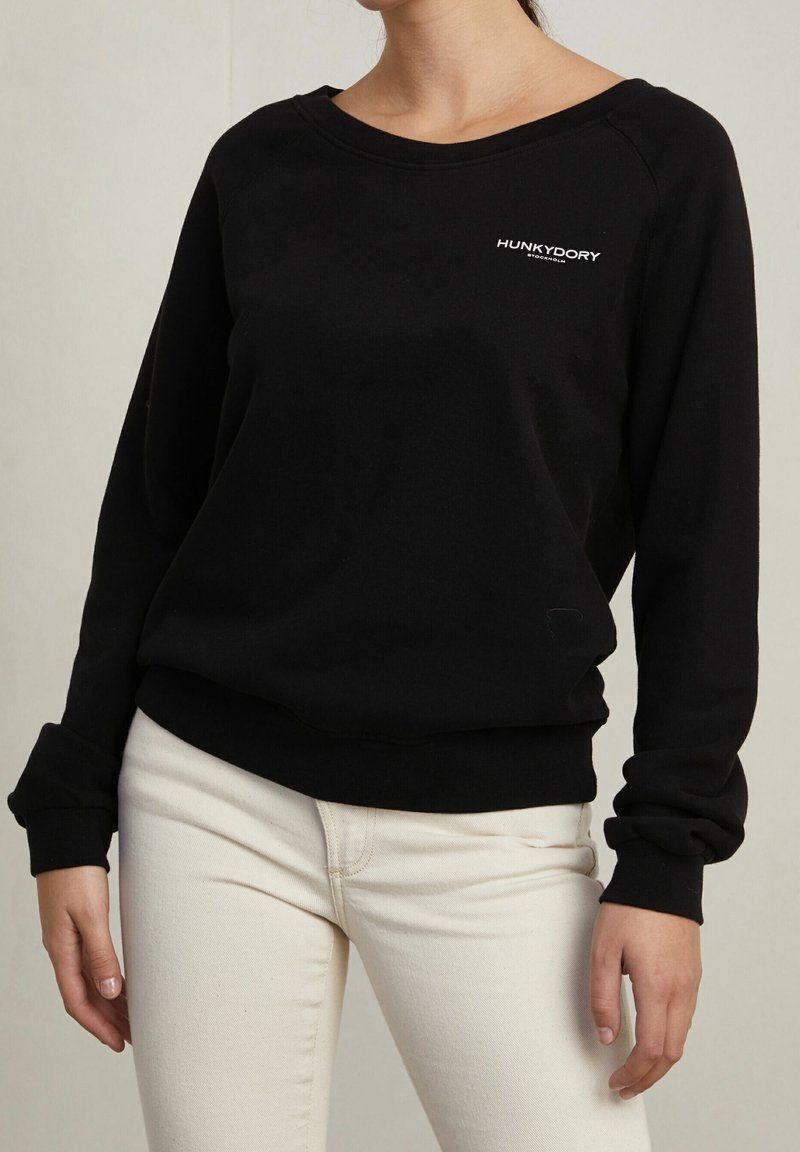 Schwarzer Sweatshirt mit rundem Halsausschnitt und langen Ärmeln. Mit einem kleinen weißen "HUNKYDORY"-Logo auf der Brust. Texturiert.