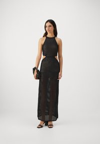 Robe maxi en mesh noir avec un col halter, des découpes sur les côtés et une silhouette ajustée. Modèle tenant un petit sac noir et portant des sandales à talons.