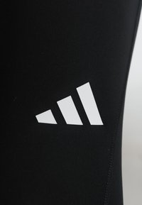 Λευκό λογότυπο με τρεις ρίγες τυπωμένο σε μαύρο ύφασμα, πιθανόν αθλητική ένδυση ή κολάν, εμφανιζόμενο με ορατή ραφή στη δεξιά πλευρά.