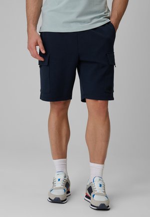 Mężczyzna w granatowych krótkich spodenkach typu cargo, białych skarpetach sportowych oraz białych, niebieskich i szarych sneakersach stoi na jednolitym szarym tle.