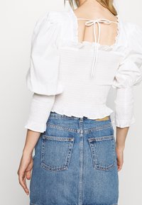 Valkoinen pörhöhiha blouse, jossa on smokkivartalo, röyhelöhelma ja takana solmimis—yksityiskohta, yhdistettynä farkkuhameeseen, jossa on takataskut.