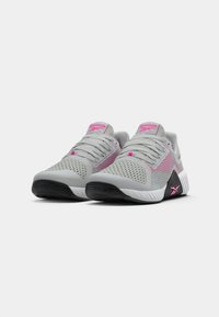 Pāris pelēku un rozā Reebok sporta apavu ar sietiņa augšdaļu un melnām zolēm, novietoti blakus uz baltas fona.