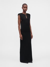Femme grande portant une longue robe noire sans manches avec des épaules rembourrées et une petite ouverture en forme de trou de serrure à l'avant.