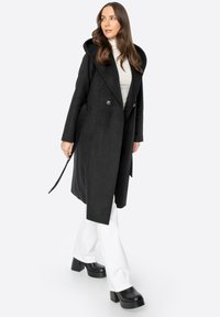 Černý kabát ze směsi vlny s velkou kapucí, s dvojitým zapínáním na knoflíky a hladkou texturou. Nosený přes světlý rolák a bílé kalhoty.