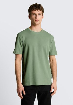 Young man with curly hair wearing a plain light green short-sleeve shirt and black pants, standing against a neutral background.