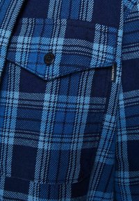 Niebieska koszula w kratę, zawierająca mieszankę jasnych i ciemnych odcieni niebieskiego, z zapinaną na guziki kieszenią na klatce piersiowej oraz fakturą materiału typową dla flaneli.