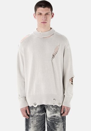 Joven con un suéter de punto beige desgastado con agujeros, combinado con jeans rasgados en blanco y negro, mirando hacia adelante con una expresión neutral.