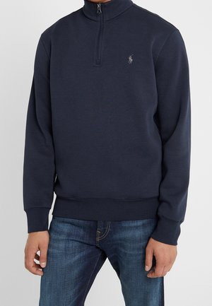 Marineblå quarter-zip pullover med logo på brystet, båret med blå jeans, vist fra midten af torsoen til hænderne mod en lys baggrund.