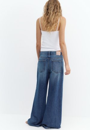 Ženska z dolgimi valovitimi lasmi, oblečena v belo majico brez rokavov in široke modre kavbojke, stoji bosa s hrbtom obrnjena proti enobarvni podlagi.