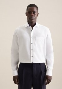 Mand stående vendt fremad iført en hvid skjorte med sorte knapper og mørke bukser mod en ensfarvet baggrund.