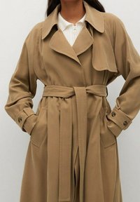 Beige Trenchcoat mit weitem Kragen, Bindegürtel und geknöpften Manschetten. Verfügt über Seitentaschen und eine glatte Stofftextur.