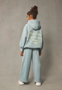 Vaaleansininen, ylikokoinen huppari, jossa teksti selässä, yhdistetty vastaaviin leveälahkeisiin housuihin. Materiaali vaikuttaa pehmeältä ja teksturoidulta. Valkoiset lenkkarit.