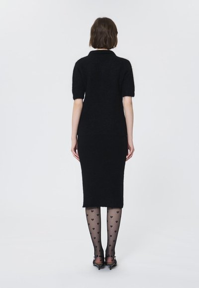 Rochie tricotată neagră, cu mâneci scurte, design ajustat, până la genunchi. Ciorapi negri transparenți cu modele în formă de inimă și tocuri negre. Fundal simplu.