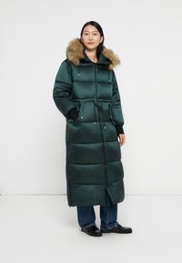Abrigo acolchado largo de color verde oscuro con capucha de piel sintética, cintura con cordón y grandes bolsillos. Material suave y brillante con secciones acolchadas.
