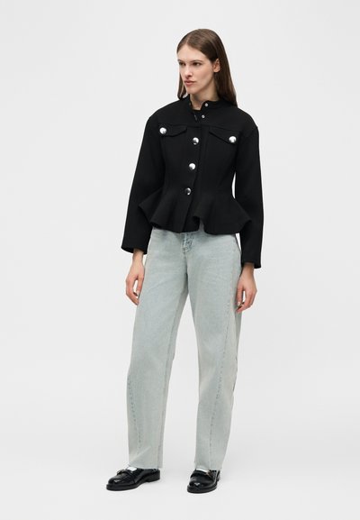 Γυναίκα που φοράει μαύρο σακάκι με κουμπιά και πεπλό, ανοιχτό μπλε τζιν με ίσια παντελόνια και μαύρες μοκασίνες, στέκεται μπροστά σε απλό φόντο.