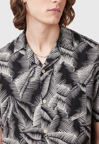 Short-sleeve black and white shirt with a leaf pattern, featuring a button-up front and a chest pocket. Made of lightweight fabric.