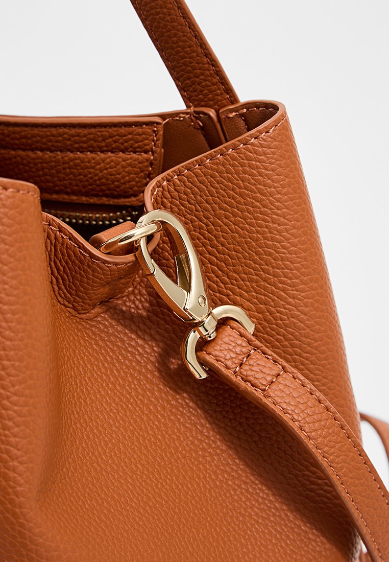 Sac à main en cuir brun texturé avec un fermoir en métal doré et une bandoulière. Présente une ouverture supérieure avec une forme douce et déstructurée.