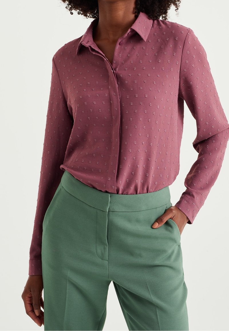 Burgundininen paita, jossa on tekstuuripainatus, napillinen kaulus ja pitkät hihat, yhdistettynä korkeavyötäröisiin vihreisiin housuihin, sileää kangasta.