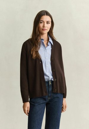 Vrouw met lang bruin haar die een bruine cardigan met knopen draagt over een blauw gestreept overhemd en donkerblauwe spijkerbroek, staand tegen een neutrale achtergrond.