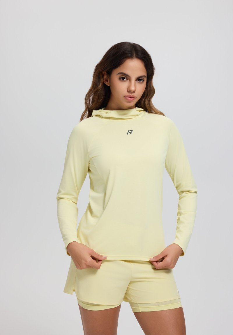 Femme portant un sweat à capuche à manches longues jaune pâle et un short assorti, debout devant un fond clair uni, tenant l'ourlet du sweat.
