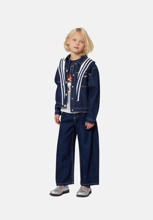 Veste en denim avec des accents blancs et un pantalon en denim à jambe large. La tenue comprend un t-shirt graphique en dessous. Des chaussures argentées complètent le look.