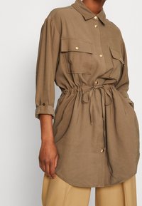 Khaki košilová košile s dvěma náprsními kapsami, se stáhnutým pasem, ohrnovacími rukávy a zaobleným spodním lemem, vyrobená z hladké, lehké tkaniny.
