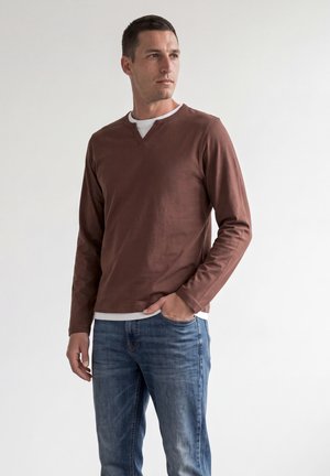 Uomo che indossa una camicia marrone a maniche lunghe sopra una canottiera bianca e jeans blu, in piedi con una mano in tasca, guardando di lato.