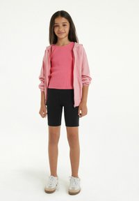 Sweat à zip rose clair, haut rose à manches courtes côtelé, shorts de vélo noirs, et baskets blanches avec des accents marron. Textures lisses, coupe décontractée.